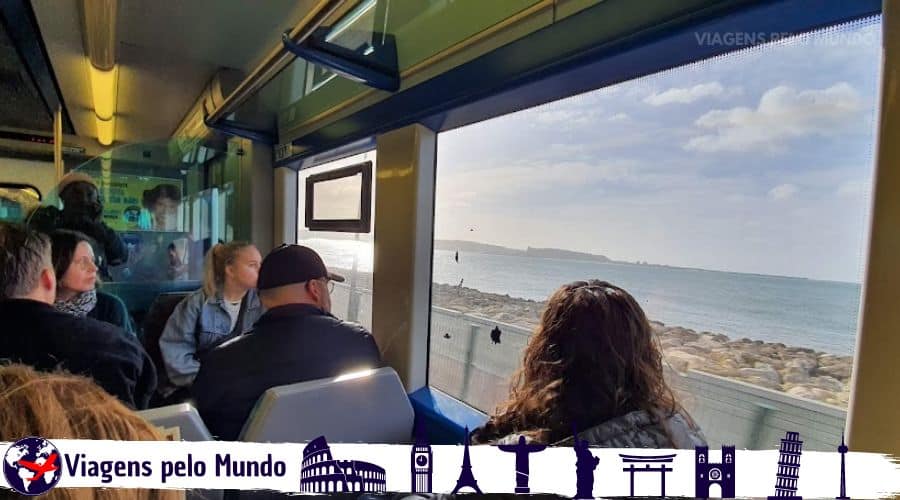 Trem que conecta Lisboa à Cascais. Pessoas sentadas olhando para janela do trem que está passando bem próximo do Oceano Atlântico