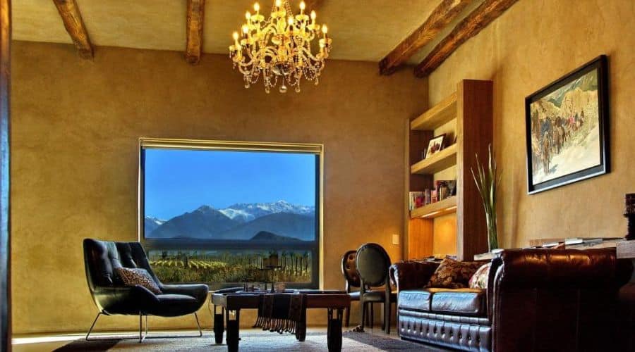 Alpasión Lodge: Sala de estar luxuosa com teto de vigas de madeira, candelabro pendente e uma grande janela com vista para as montanhas. A sala é decorada com sofás de couro, poltronas confortáveis e estantes embutidas, criando um ambiente elegante e aconchegante.