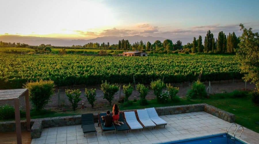 Hotéis vinícolas em Mendoza: Posada Mawida tem uma vista deslumbrante de um vinhedo ao entardecer, com montanhas ao fundo. Em primeiro plano, um casal está sentado em espreguiçadeiras próximas a uma piscina, apreciando a paisagem. O céu está colorido com tons suaves de laranja e rosa, criando um cenário romântico e tranquilo.