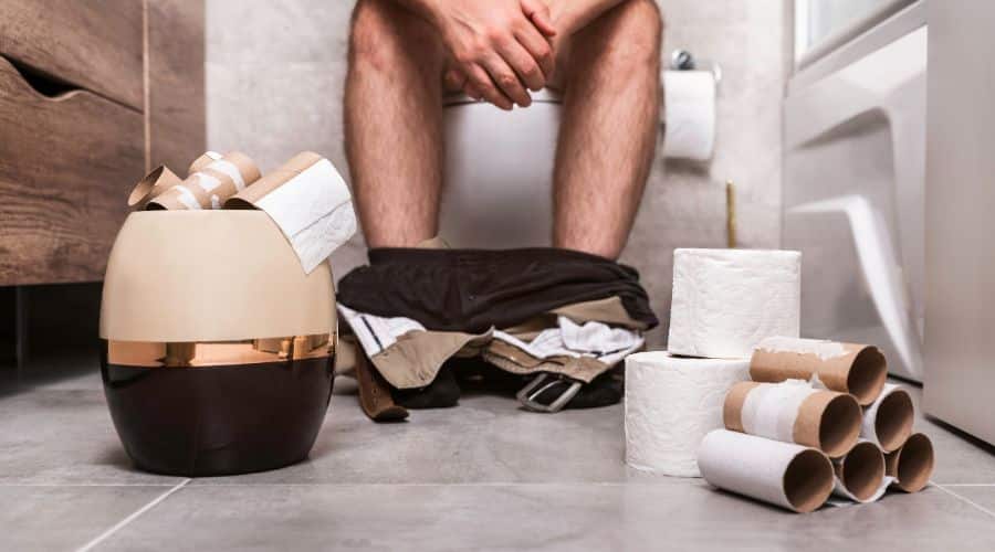 Homem sentado em um vaso sanitário com diversos rolos de papel higiênico 