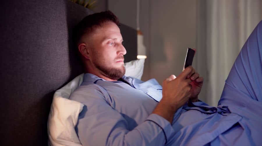 Homem deitado na cama de pijama com o celular na mão