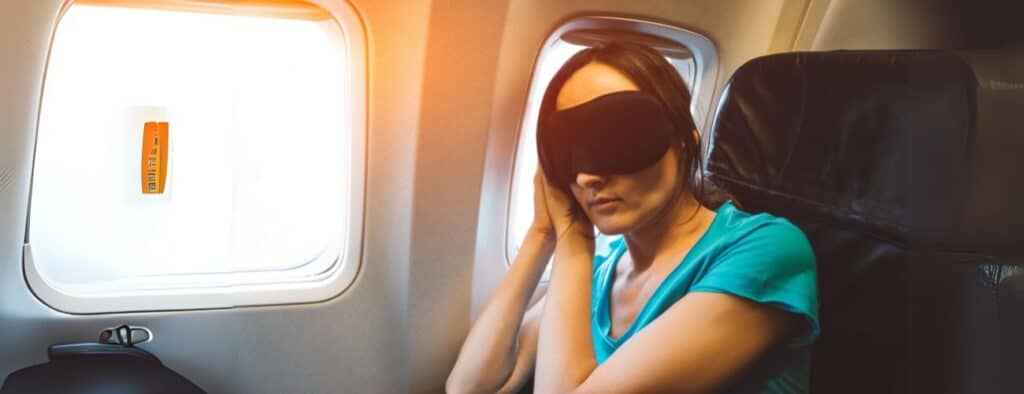 Jet Lag: Moça sentada na poltrona de avião com máscara para dormir com a janela aberta e com dificuldade.