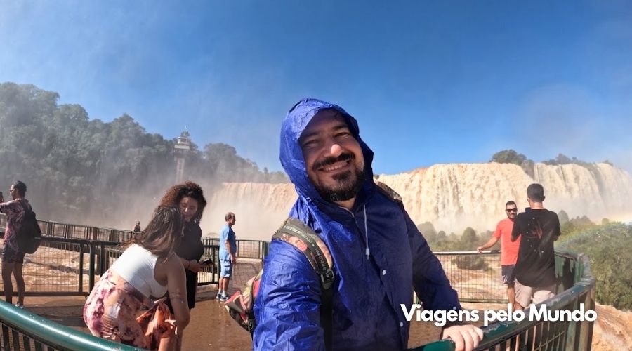 Eu com capa de chuva na plataforma e ao fundo as quedas de água das cataratas do iguaçu.